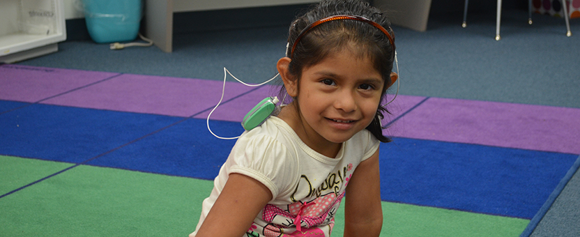 Young girl with hearing aids smiles while sitting on a classroom rug