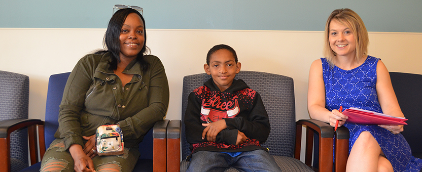 Adolescent boy and his mother smile while sitting in a clinic waiting room with their DSCC Care Coordinator