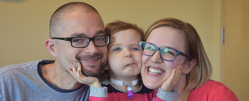 Young girl with tracheostomy tube smiles while posing between her mom and dad and holding their faces