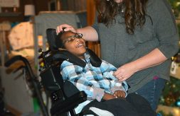 A boy with medical complexity sits in his wheelchair and smiles as his mother caresses his head