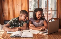 A brother and sister look at a laptop screen together while doing homework