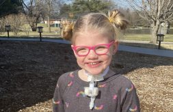 Home Care participant Willa sits outside with a big smile. She is wearing pink glasses and has a tracheostomy