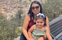 Current DSCC staff member and former participant Lisette Rios holds her young daughter in her lap as they sit on an outdoor ledge overlooking trees, hills and a city